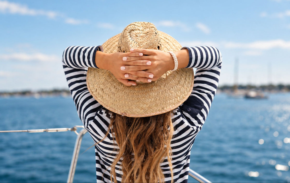 A woman on a boat seen from behind, wearing a black and white striped long-sleeve top and a wide-brimmed straw hat, with her hands behind her head, overlooking a sunny blue sea.